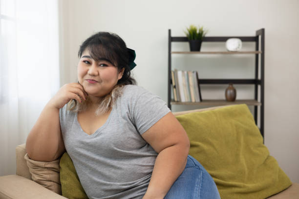 Happy women's plus sitting on sofa in living room. Body positive plus size female lives happily and is proud of herself. Happy women's plus sitting on sofa in living room. Body positive plus size female lives happily and is proud of herself. plus size photos stock pictures, royalty-free photos & images