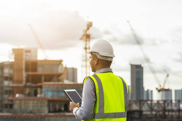istock Asian engineer handsome man or architect use tablet with white safety helmet in city construction site . Standing on rooftop building construction at capital. 1356792410