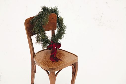 Merry Christmas! Christmas wreath on rustic chair in snowy winter field. Winter holidays in countryside. Space for text. Stylish xmas wreath with pine branches and red bow hanging on wooden chair