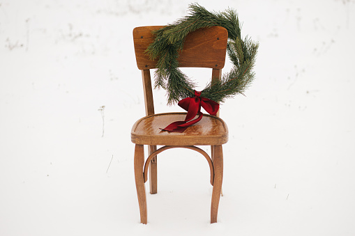Merry Christmas! Christmas wreath on rustic chair in snowy winter field. Winter holidays in countryside. Stylish xmas wreath with pine branches and red bow hanging on wooden chair. Atmospheric time
