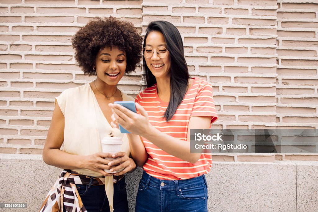 Best friends enjoying in Madrid, using smartphone and having fun Two beautiful friends on a journey in Madrid drinking coffee and using smartphone on the move. Friendship Stock Photo
