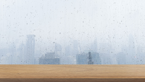 empty wooden table top in foreground with wet transparent window glass, highrise buildings view on background. counter bar foreground can be used for display or montage products.