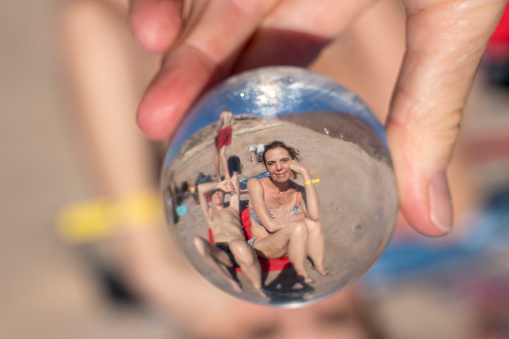 Hand holding crystal ball that reflect people enjoying on sandy beach.