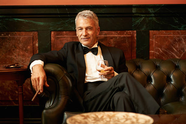 Suave man in a cigar lounge Portrait of a sophisticated senior man sitting on couch holding cigar and glass of brandy rich man stock pictures, royalty-free photos & images