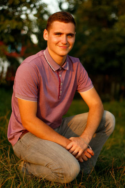 幸せな若者。夏の日に自然の背景に屋外に座っている紫色のポロシャツで笑顔の若いブルネットの男の肖像画。20年代の男。ミレニアル世代。外。笑顔の人 - 20s 20 25 years beauty beautiful ストックフォトと画像