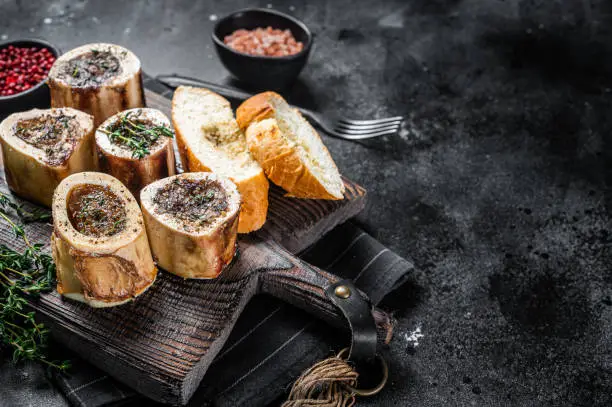 Roast marrow calf bones on wooden board with bread and herbs. Black background. Top view. Copy space.