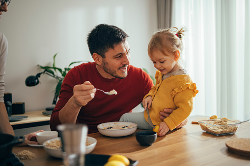 Father and small daughter are sitting at a kitchen table in the morning and having a healthy breakfast. The man is feeding the child with cereal. There are plates with cereal on the table. The kid is holding a lemon and a knife. They are both smiling. Mother is standing next to them.