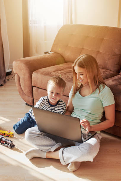 Young woman with child working on laptop Close up portrait of young blond caucasian woman with little son. They are sitting on the floor at home. Mother is working on computer and bored child is leaning on her Distracted stock pictures, royalty-free photos & images