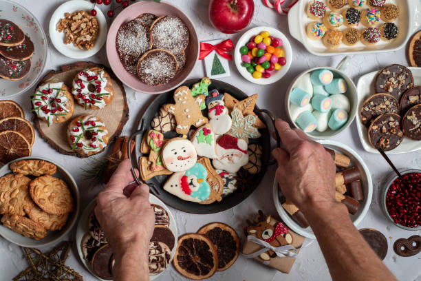 Christmas gingerbread cookies in a metal bowl in men's hands on a table full of Christmas sweets, pralines, mini kuglof, manchmelou and colorful Christmas candies, chocolates, fruits, biscuits with Christmas decorations. Person serving festive treats first person view