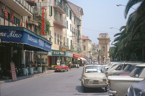 Savona Province, Liguria, Italy, 1969. Street scene with restaurants, pedestrians, vehicles, buildings and a promenade with palm trees in Savona.