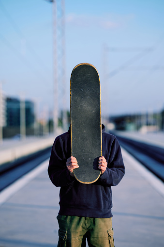 A teenage boy is standing at the train station and holding a skateboard.