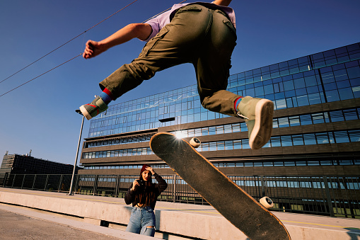 A teenage boy performs tricks on a skateboard while a girl takes photos of him.