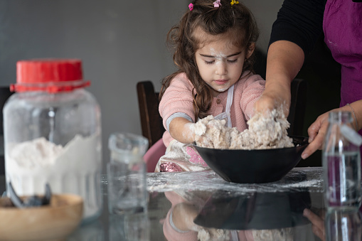 Photo of 3,5 years old little girl helping mother in cooking. The motel is wearing a pink sweater. She in sittin on table. Shot indoor with a full frame mirrorless camera.