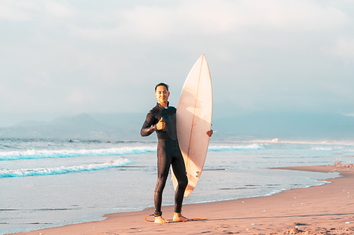 surfer on the beach smiling with a surfboard and ocean background, thumb up