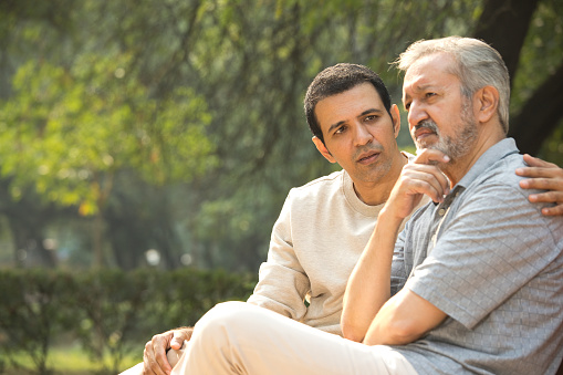 Son comforting thoughtful old father sitting on bench at park