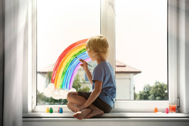 маленький мальчик рисует радугу на окне в помещении. концепция «оставайтесь дома» - primary care стоковые фото и изображения