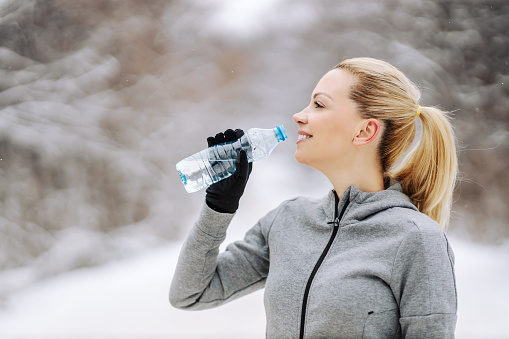 Happy sportswoman standing in nature at snowy winter day, taking a break and drinking fresh water. Healthy habits, refreshment