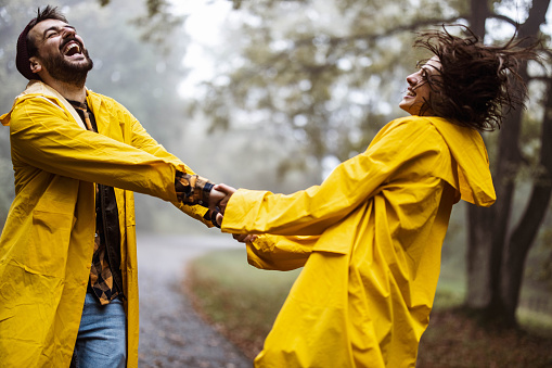 Happy couple in raincoats having fun while holding hands and spinning in nature.