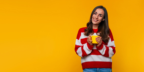 Banner photo of cute happy young brunette woman with yellow cup of hot tea or coffee in hands isolated on yellow background