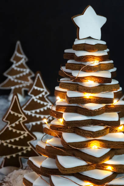 Close-up image Christmas tree, stacked gingerbread star cookies iced with white royal icing on wooden cake stand, decorated by illuminated fairy lights, conifer forest scene with Christmas tree biscuits, artificial snow, black background