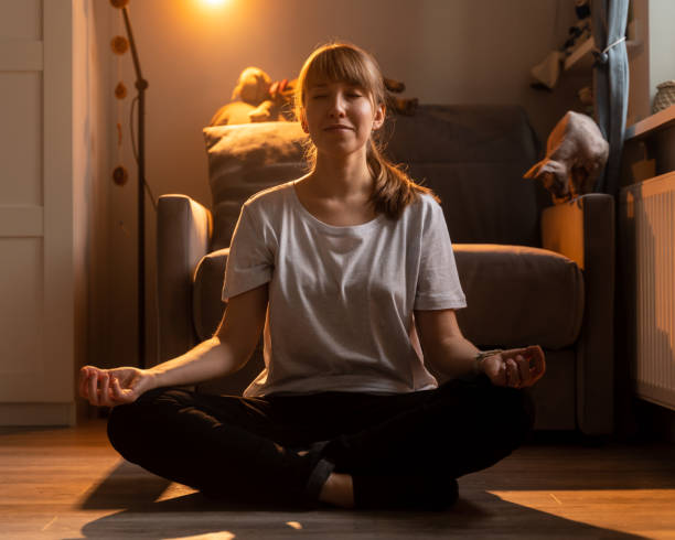 Young smiling female meditating in lotus pose. Doing yoga at home in cozy evening stock photo