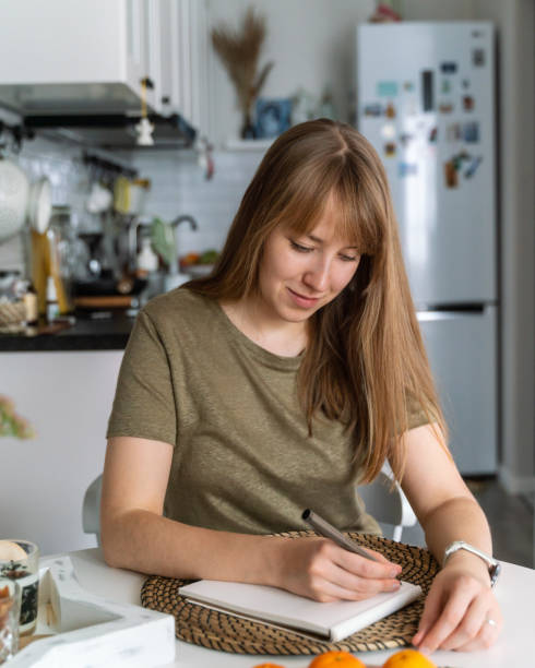 istock Young female making notes in notebook sitting in kitchen. Planning buying. Making list of products 1356934578