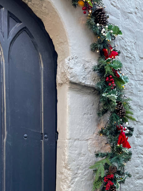 istock Image of blue, arched, wood front door with panels in white stone wall, surrounded by artificial pine needle garland archway illuminated white fairy lights, decorated with red berries, pine cones, holly leaves, white mistletoe berries and red velvet bows 1356944439
