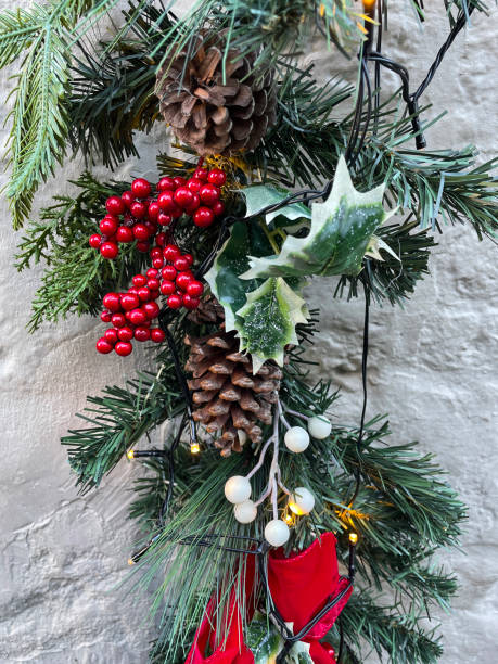 istock Close-up image of white stone wall with artificial pine needle garland archway illuminated white fairy lights, decorated with red berries, pine cones, holly leaves, white mistletoe berries and red velvet bows 1356944444