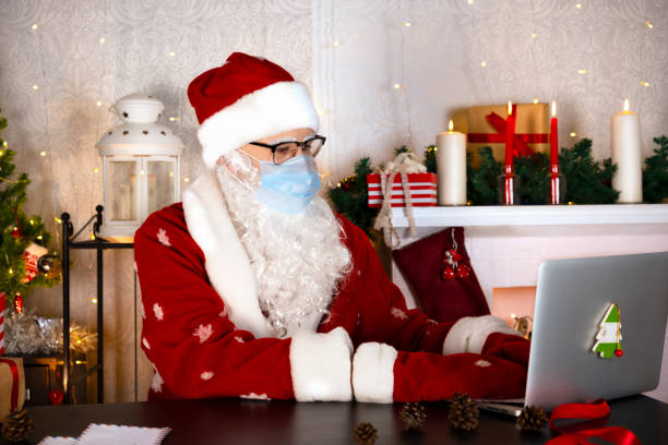 istock Santa Claus in blue medical protective mask types on laptop computer at his workshop home. 1356947449