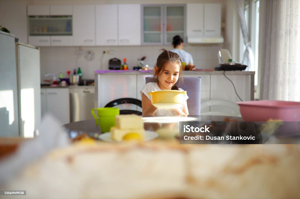 Happy girl, sift the flour onto dining table int he kitchen Adorable and cute girl, sift the flour at the kitchen, while her mother working behind her 35-39 Years Stock Photo