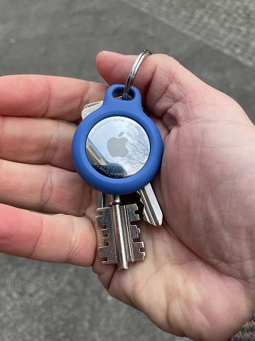 A male hand is holding a set of keys with an Apple AirTag attached to the key ring  with a Belkin adapter. Outdoors, daytime.