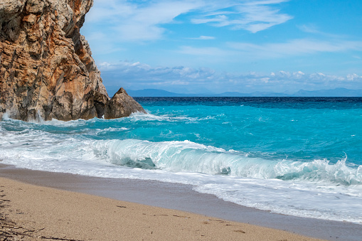Azure sunny stormy waves with white foam breaking rocky coast. Sandy beach with blue sky on Lefkada island in Greece. Summer nature travel to Ionian Sea
