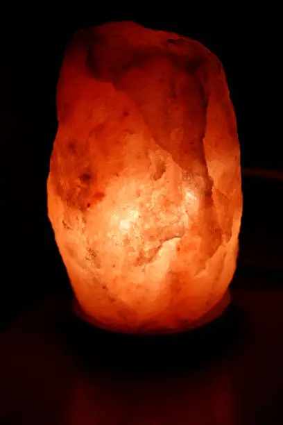 Large lit glowing dark pink Himalayan salt lamp on a wood base closeup. In a dark room is shining a glowing half a foot or so high, oblong, dark pink Himalayan salt lamp on a round wood base close-up