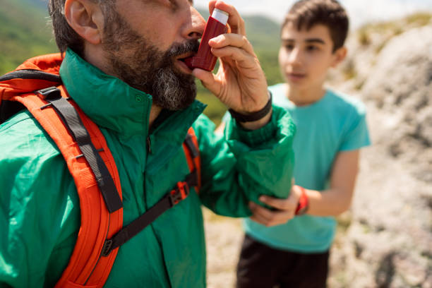 Man inhaling himself with asthma inhaler, while his worried son giving him support, during their hike stock photo