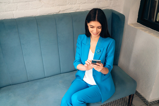 Elegant young Caucasian businesswoman in blue suit, using mobile phone while shopping online, from the office