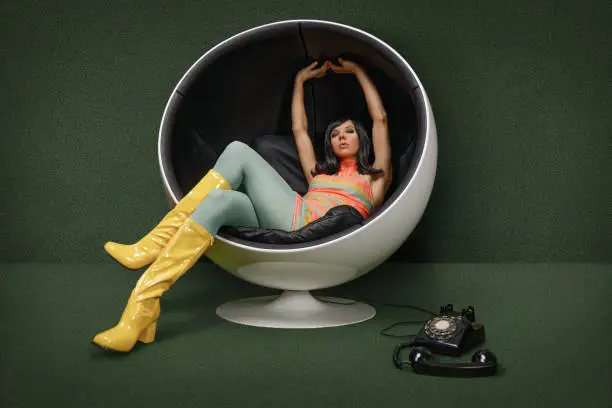 Photo of Young woman from the 60s sitting in a vintage ball chair, her rotary dial telephone of the hook at her feet