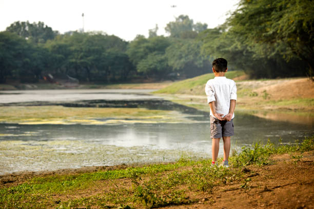 istock Boy standing with hands behind back at riverbank 1356996800