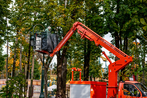 Kharkiv, Ukraine - October 20, 2020: Worker changes light bulbs in street lamps, standing on an aerial platform, in Maxim Gorky Central Park for Culture and Recreation (Kharkiv). Man works in the autumn city garden