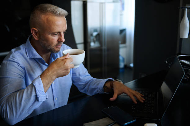 istock Charming middle aged European man relaxing, drinking coffee while working on laptop, sitting at table at home office during lockdown 1357012323