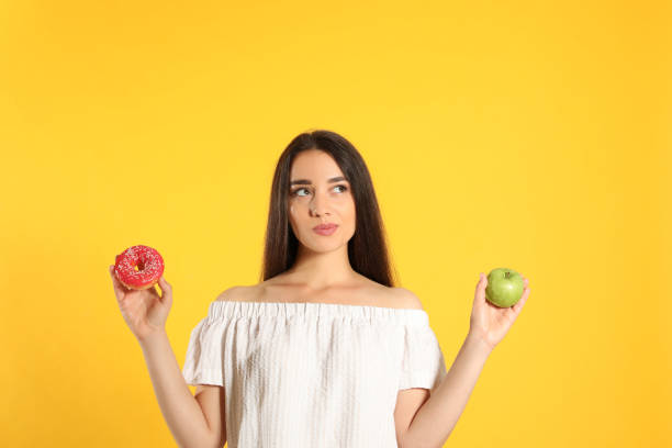 istock Doubtful woman choosing between apple and doughnut on yellow background 1357013518