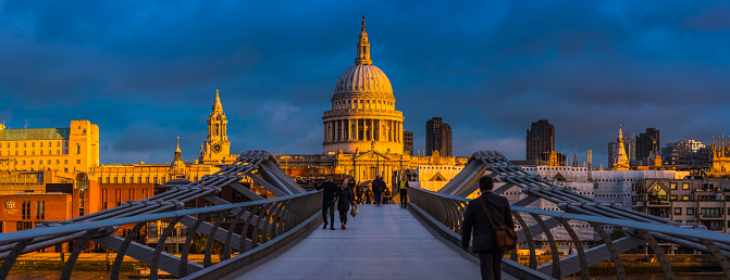 Golden light of sunrise illuminating the futuristic walkway of the Millennium Bridge crossing the River Thames at Bankside towards the iconic dome of St. Paul’s Cathedral in the City of London.