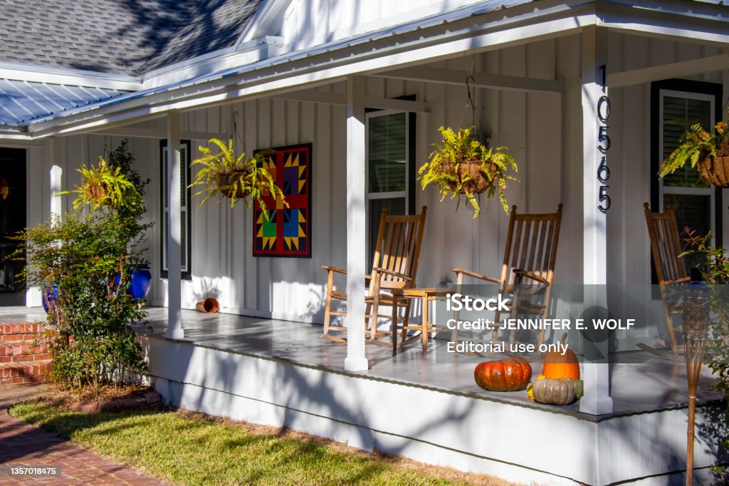Hanging ferns, wooden rocking chairs, colorful art, and a collection of pumpkins create an inviting seasonal look for the front porch of a renovated wood-frame house in historic White Springs, Florida White Springs, Florida - November 24, 2021: Hanging ferns, wooden rocking chairs, colorful art, and a collection of pumpkins create an inviting seasonal look for the front porch of a renovated wood-frame house in the historic center of town. Porch Stock Photo