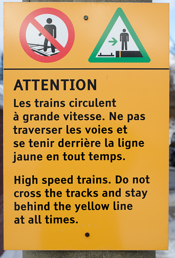 Bilingual warning sign at train station stating to be careful to stay behind the line and not to cross the tracks