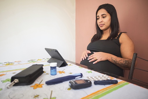 Portrait of a young Latin pregnant woman at home, using her tablet on a video call with a doctor. She has a glaucometer and some diabetes medicine on the table, due to a gestational diabetes condition.