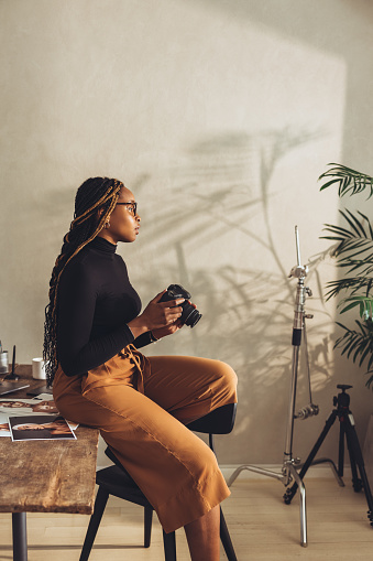 Female artist holding a dslr camera in her home office. Creative young photographer looking thoughtful while sitting on her desk. Freelancer working on a new project in her workspace.