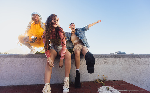 Energetic female friends enjoying themselves outdoors. Three happy friends smiling while hanging out on a wall in the city. Cheerful female youngsters having fun together.