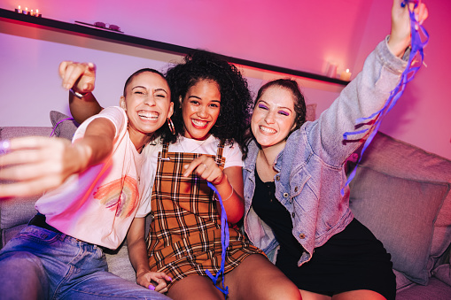 Three girlfriends having a good time at a house party. Vibrant young women celebrating while sitting on a couch in bright neon light. Cheerful female friends having fun together on the weekend.