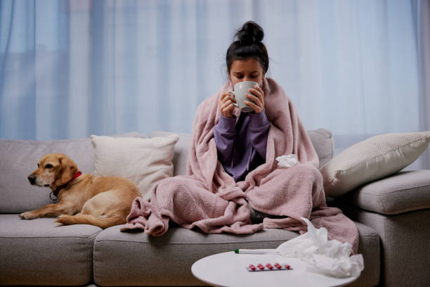 어린 아픈 여성은 작은 개와 함께 소파에 누워 뜨거운 차를 마시고 계절 독감이나 감기로 고통받고 있습니다. 아픈 소녀는 집에서 인플루엔자로 건강에 해로운 느낌 - illness pets symptoms blanket 뉴스 사진 이미지