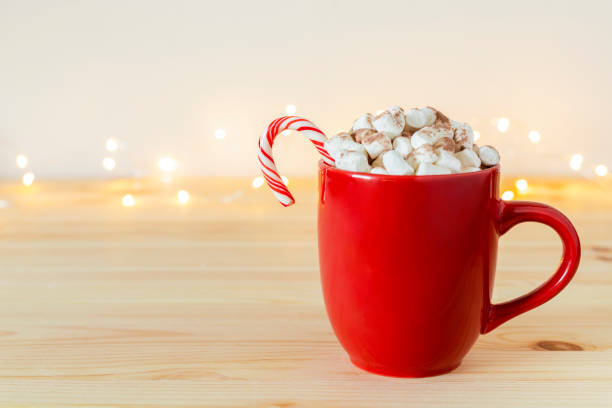 istock Cocoa in red mug with marshmallows and candy cane on table against white wall background with holiday lights. 1357054868