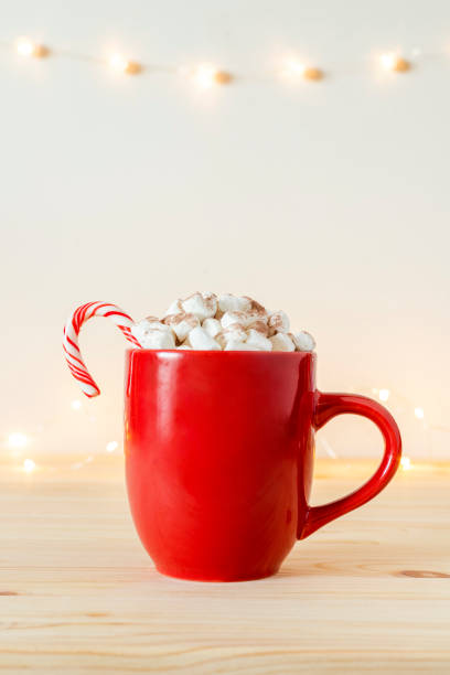 istock Hot chocolate or cacao in red mug with small marshmallows and candy cane on table with holiday lights on wall. 1357054869